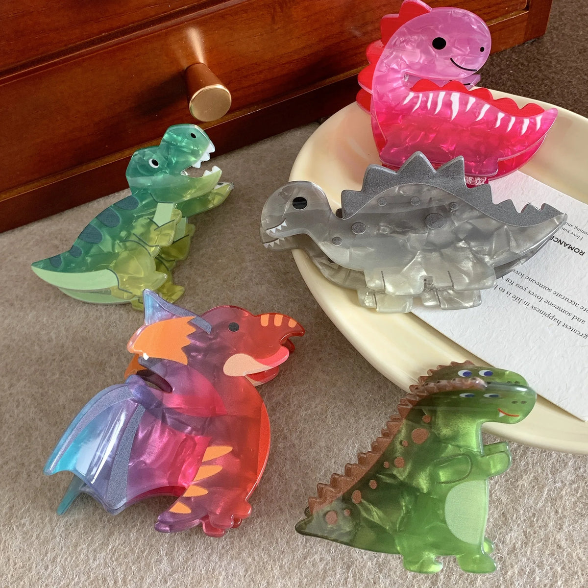 Five translucent dinosaur-shaped hair claw clips in various colors and designs, including green, pink, gray, orange, and blue, arranged on a beige surface next to a wooden drawer and a cream-colored dish.