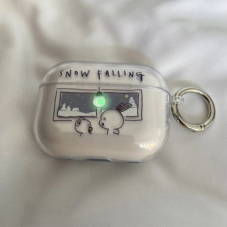 A clear protective case for an AirPods charging case, featuring a cute cartoon illustration of a rabbit and a bird looking at a snowy window with the text 'SNOW FALLING' above. The case has a silver metal key ring attached and is shown on a white fabric background.