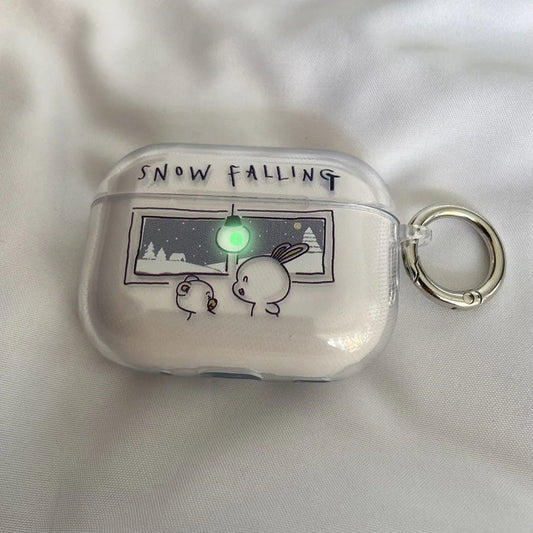 A clear protective case for an AirPods charging case, featuring a cute cartoon illustration of a rabbit and a bird looking at a snowy window with the text 'SNOW FALLING' above. The case has a silver metal key ring attached and is shown on a white fabric background.