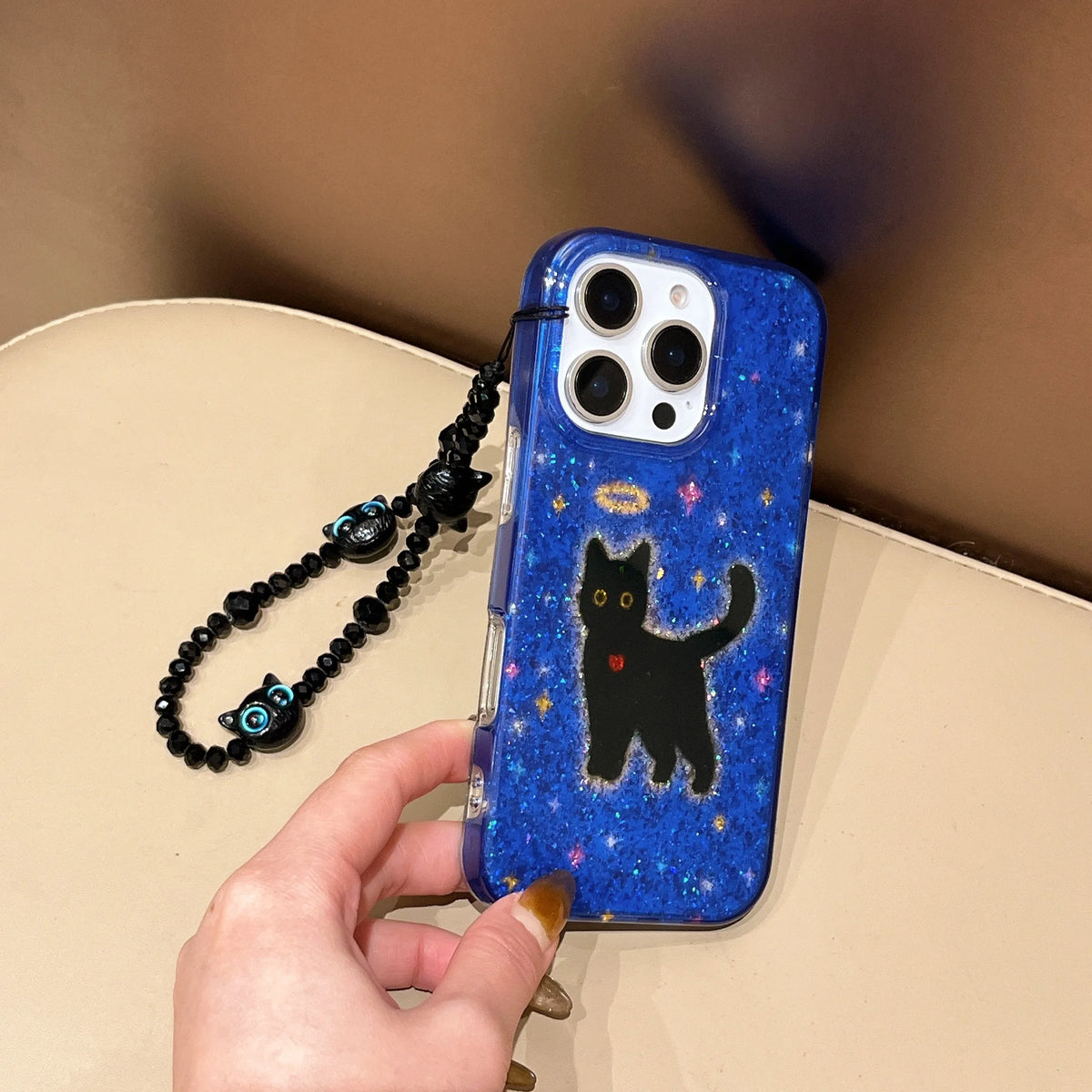 A hand holds a blue glittery phone case featuring a black cat illustration with a small gold halo and red heart, complete with a black beaded wrist strap. The phone case is shown on a beige surface against a brown background.