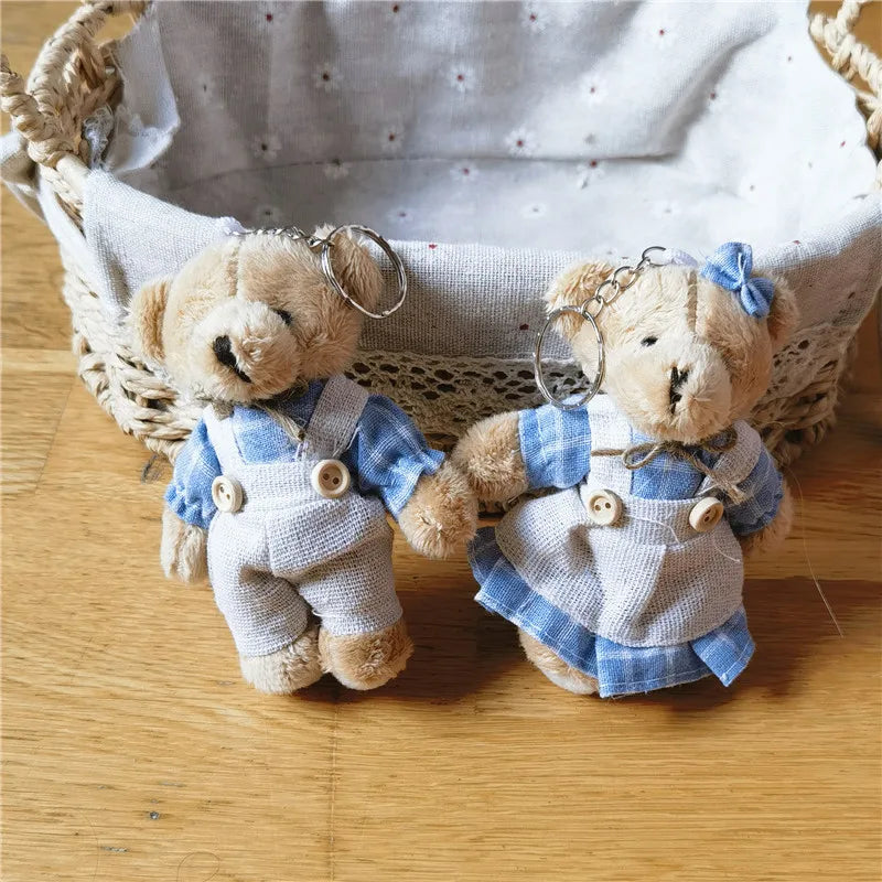 Two small teddy bear keychains dressed in blue and white outfits, standing on a wooden surface in front of a woven basket with a light fabric lining.