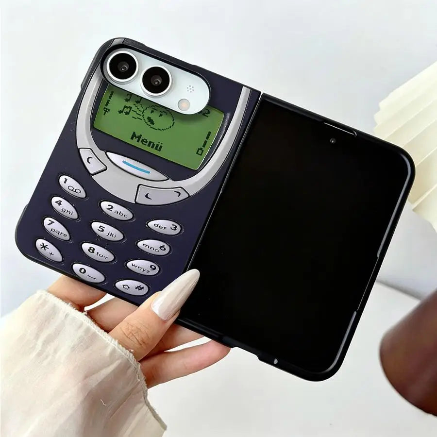 A black flip-style phone case designed to look like a retro mobile phone, held in a hand. The case features a dark navy/black body, a silver-gray border around the screen, a green display screen with a cartoon face, and white phone buttons on the front cover.