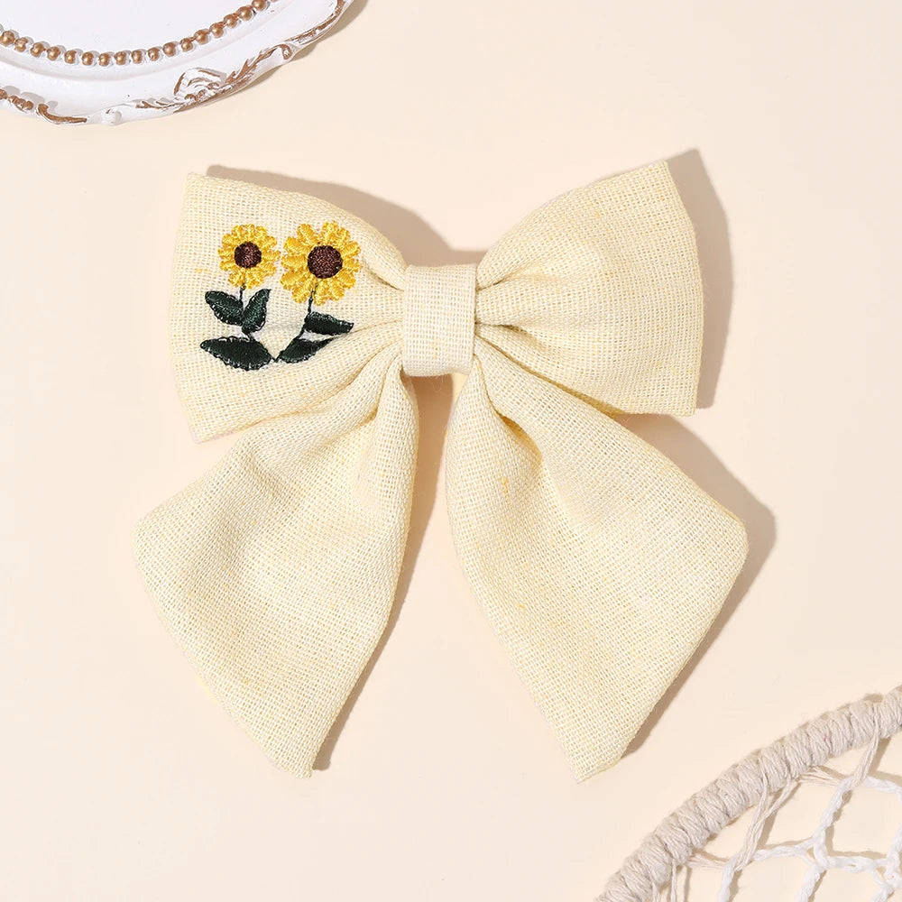 A pale cream-colored woven fabric hair bow featuring embroidered yellow sunflowers with green leaves on the left side, displayed on a warm beige background with decorative props.
