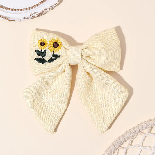 A pale cream-colored woven fabric hair bow featuring embroidered yellow sunflowers with green leaves on the left side, displayed on a warm beige background with decorative props.