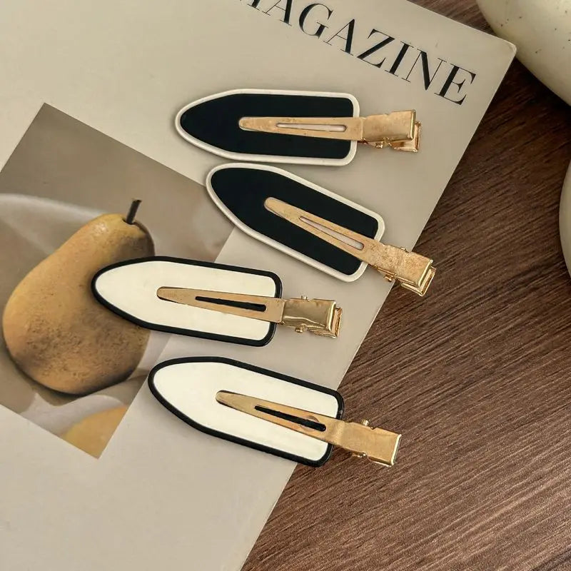 Four hair claw clips with a black and white color-blocked design and gold-tone metal clips, arranged on a magazine page over a wooden surface.