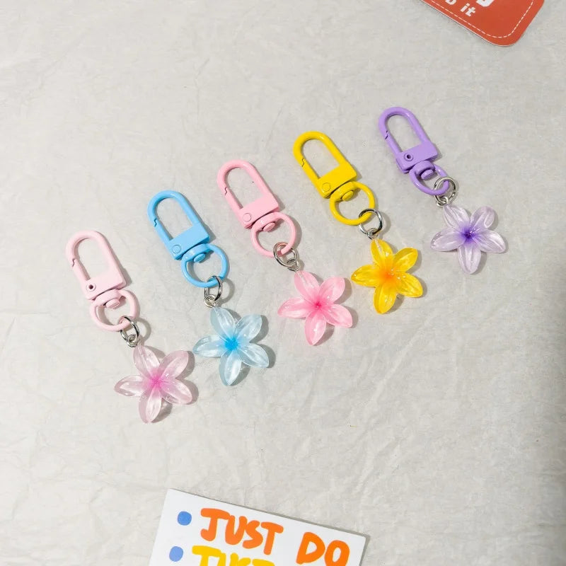 Five small plastic flower keychains in pastel pink, blue, light pink, yellow, and lavender, each attached to a metal clasp, arranged on a light textured surface.