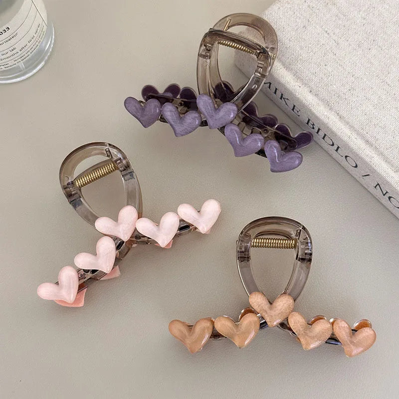 Three claw-style hair clips arranged on a light gray surface, each decorated with a row of glossy heart charms in different colors: lavender, pale pink, and nude beige. A white textured book and part of a bottle are visible as styled props.