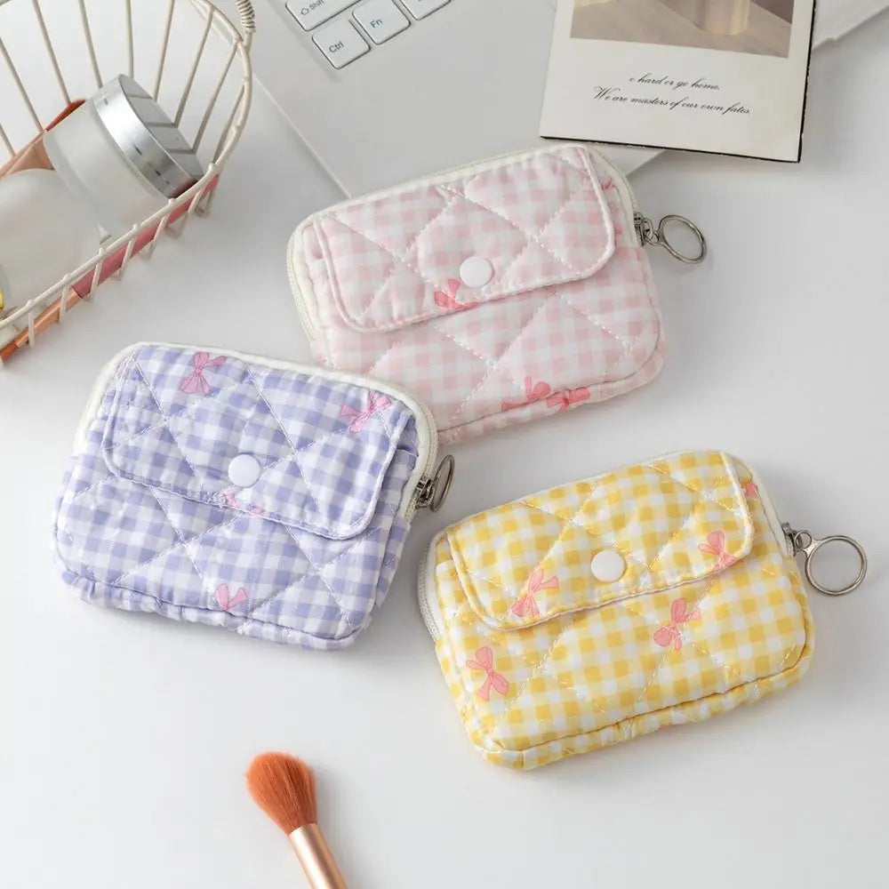 Three small quilted zip pouches with snap closures are arranged on a white surface. One is light pink gingham with tiny pink bow motifs, one is light purple gingham with tiny pink bow motifs, and one is light yellow gingham with tiny pink bow motifs.