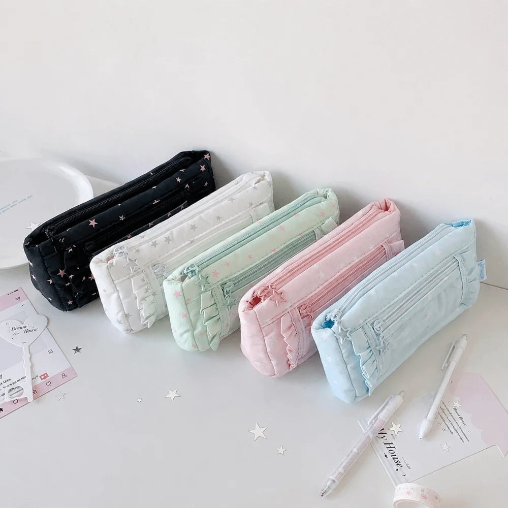 Five quilted zippered pencil cases in black, white, mint green, pink, and light blue, arranged diagonally on a white desk with stationery props.