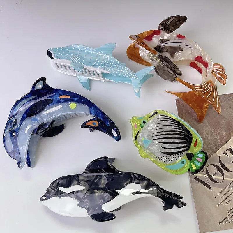 Five translucent acrylic fish and shark hair clips arranged on a white surface, with a folded Vogue magazine partially visible under one figure.