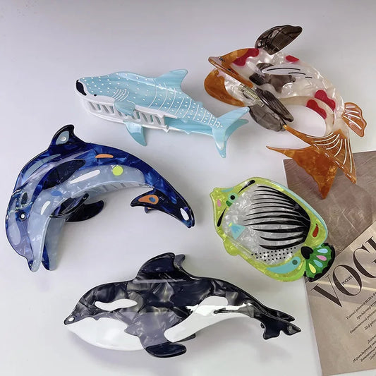 Five translucent acrylic fish and shark hair clips arranged on a white surface, with a folded Vogue magazine partially visible under one figure.