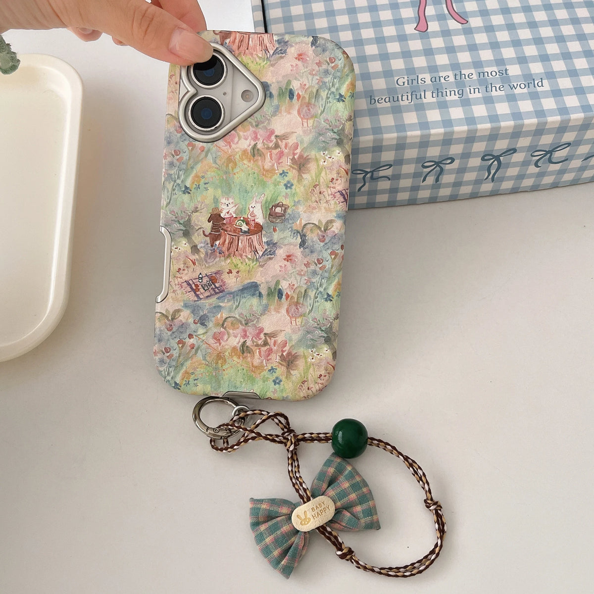 A phone case with a vintage-style watercolor illustration of a picnic scene, held by a hand. The case features a silver metal camera ring and a built-in card slot.