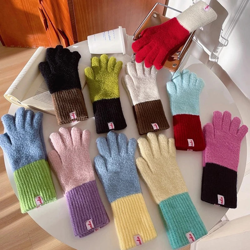 A set of color-blocked knit gloves arranged on a round white table, with one glove held in a metal cup. The gloves come in multiple colorways including black/brown, lime green/brown, cream/brown, light blue/brown, red/brown, light blue/pink, lavender/pink, yellow/teal, yellow/teal, yellow/pink, and pink/black.