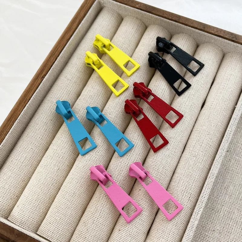 Six colorful zipper pull earrings in yellow, black, blue, red, and pink arranged inside a beige fabric-lined jewelry organizer tray.