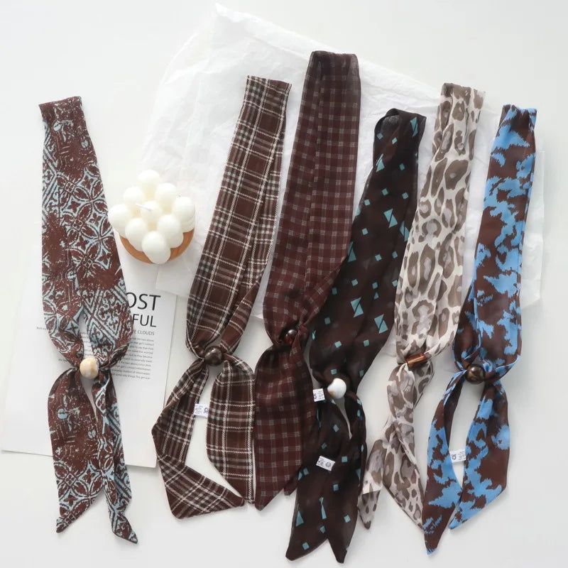 Six silk neckties with unique patterns and colors are arranged on a white surface with a white tulle bag and a small decorative candle. The neckties include a brown and white floral print, a brown and white plaid pattern, a dark brown and red plaid pattern, a dark brown and black pattern with light blue accents, a beige and brown leopard print, and a brown and blue camouflage pattern.
