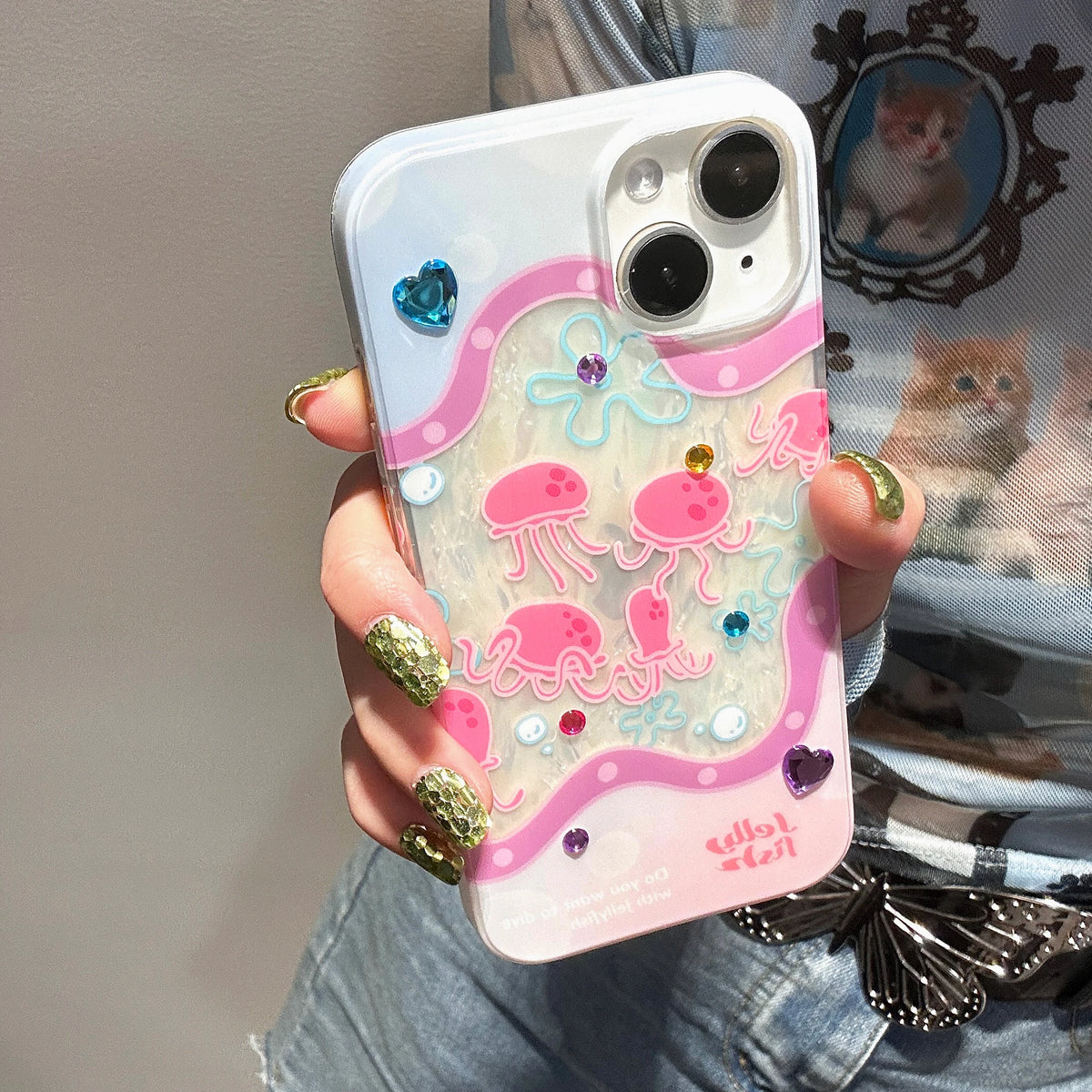 A hand with glittery green nails holds a clear phone case decorated with pink jellyfish, pink and blue wave patterns, and small rhinestones. The case has a white camera cutout and pink branding text at the bottom right.