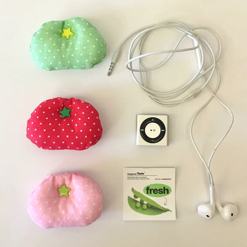 Three small fabric-covered tomato shaped objects in green, red, and pink with white polka dots and star accents are arranged on a light background beside a coiled white earphone cable, a white iPod-style music player, and a small card reading 'impressive tastes fresh'.