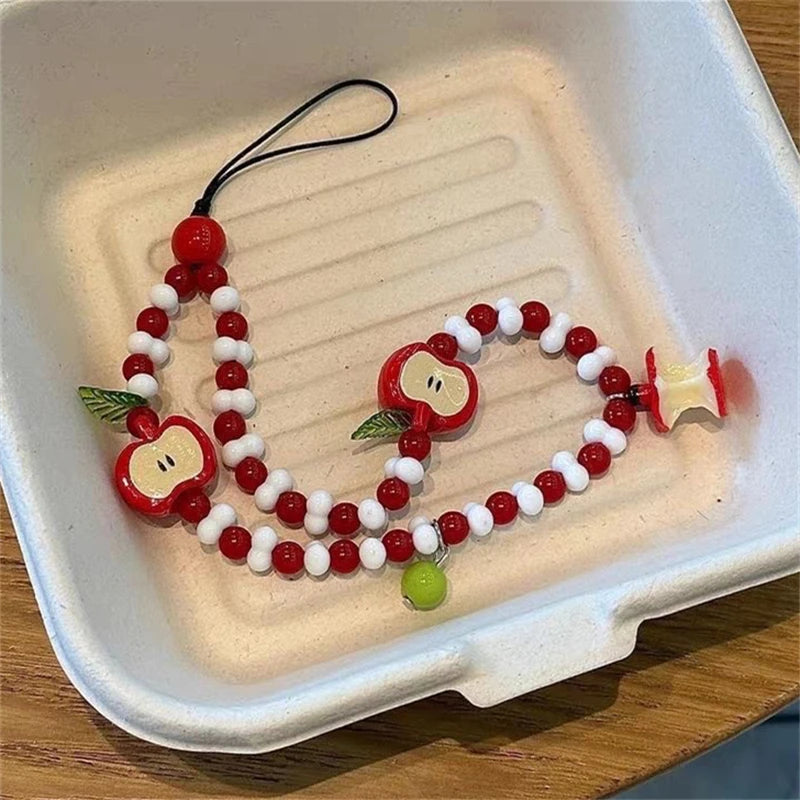 A beaded phone strap or bag charm made of red and white beads with apple-shaped charms and a small green bead, displayed inside a white tray on a wooden surface.