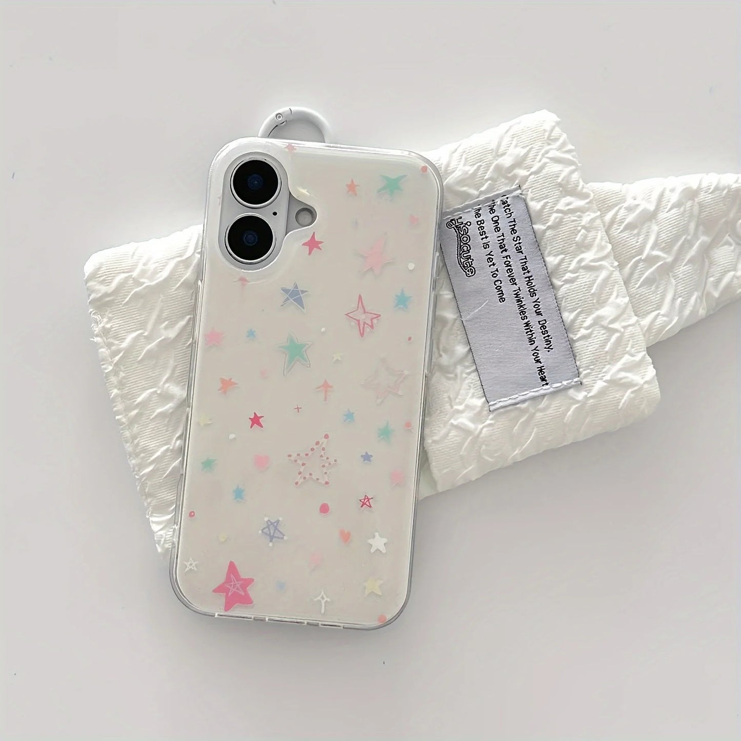 A clear phone case decorated with pastel-colored star and dot patterns, shown on a white phone and resting on a crinkled white fabric prop with a brand tag.