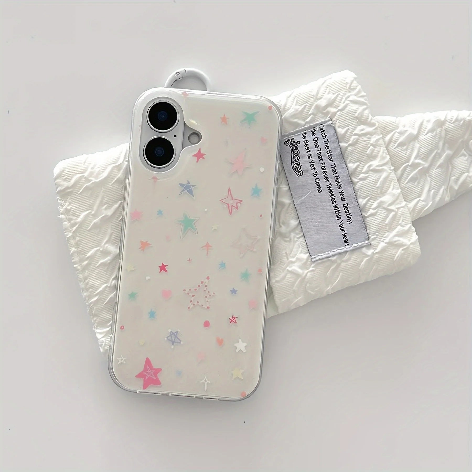 A clear phone case decorated with pastel-colored star and dot patterns, shown on a white phone and resting on a crinkled white fabric prop with a brand tag.