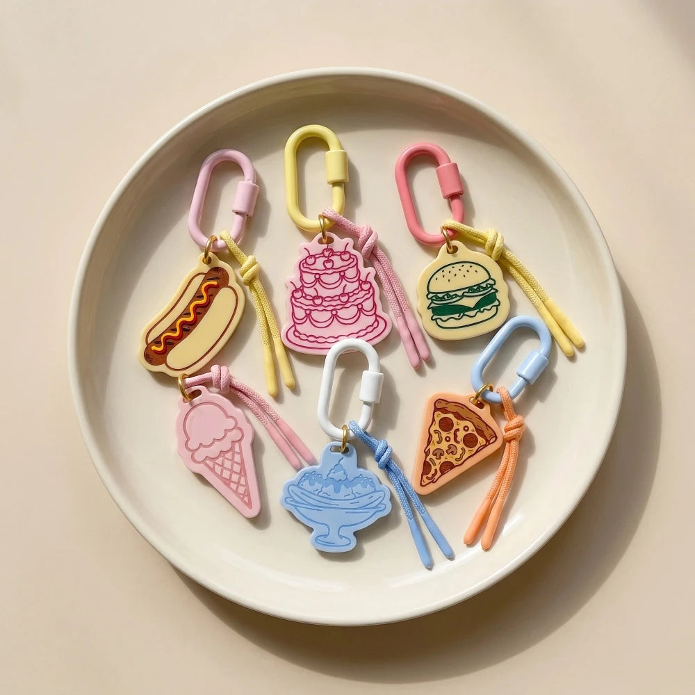 Six colorful silicone keychain charms shaped like food items (hotdog, cake, burger, ice cream cone, sundae, and pizza slice) with different colored carabiners, arranged on a round off-white plate against a warm beige background.