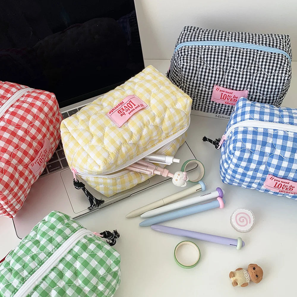 Five quilted zippered pencil cases in different gingham patterns (red, yellow, blue, green, and black-and-white) arranged on a desk with stationery items.