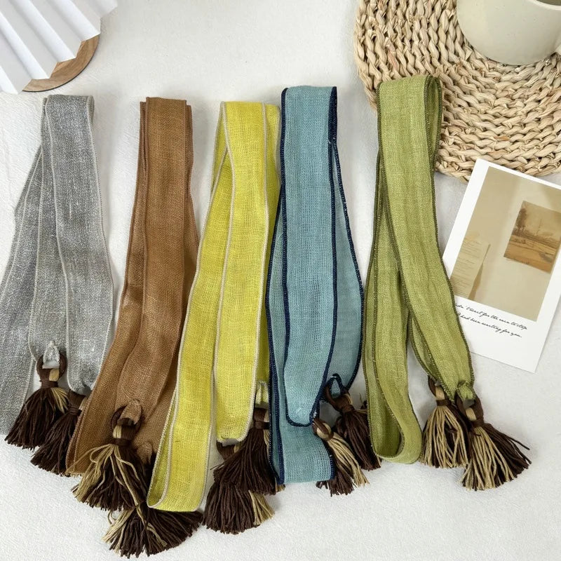 Five woven tassels in different colors are laid out on a light surface: gray, tan, mustard yellow, dusty blue, and olive green. Each tassel has a long narrow woven strap with a dark edge and a brown tassel at the end.