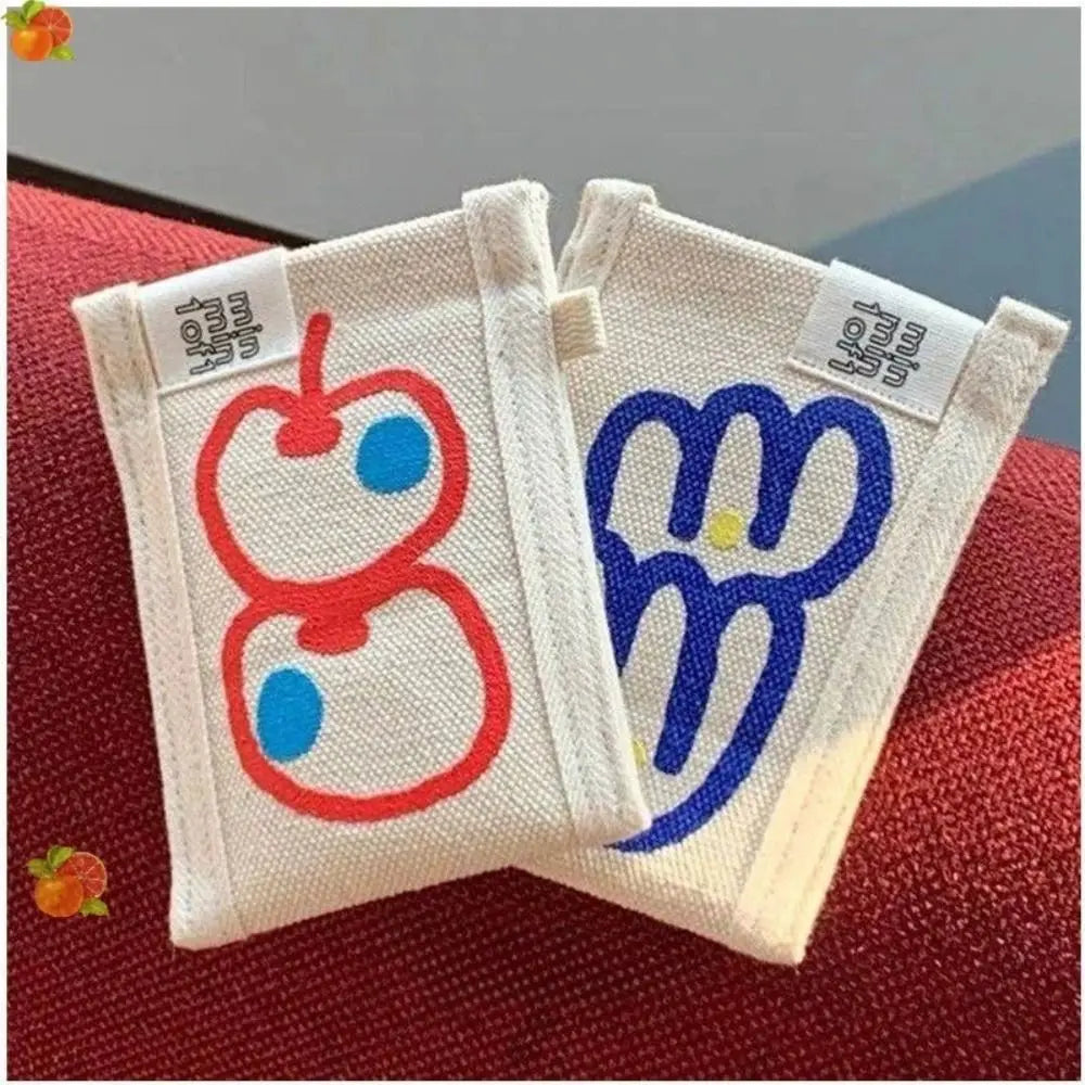 Two small white fabric pouches or bags with embroidered cartoon motifs, one in red and one in blue, resting on a red textured surface.