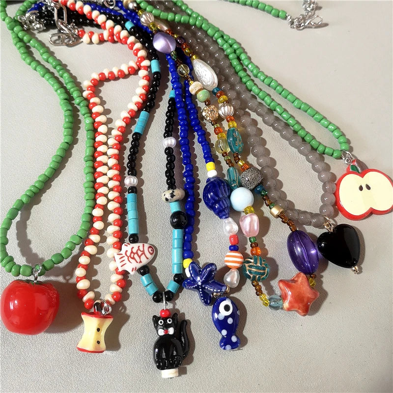 A group of colorful beaded necklaces with various charms, including a red apple, a striped cone, a black cat, a blue butterfly, a black evil eye, a purple heart, and a red-and-white apple slice, arranged on a light gray surface.