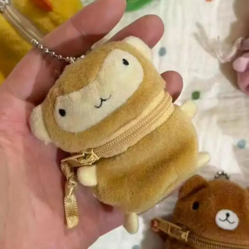 A small plush monkey keychain or coin purse held in a person's hand. The plush is tan and cream with embroidered facial features, a gold zipper around the neck, and a gold ball chain attached.