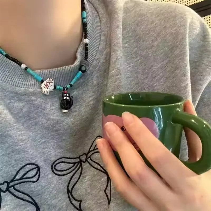 A close-up lifestyle shot of a person wearing a gray sweatshirt and holding a green ceramic mug. The person is also wearing a beaded necklace with turquoise, black, and white beads, featuring a small black cat pendant and a white cat charm.