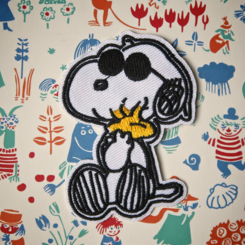 An embroidered patch featuring the character Snoopy from the Peanuts comic strip, wearing sunglasses and hugging Woodstock, on a background with various colorful characters and objects.
