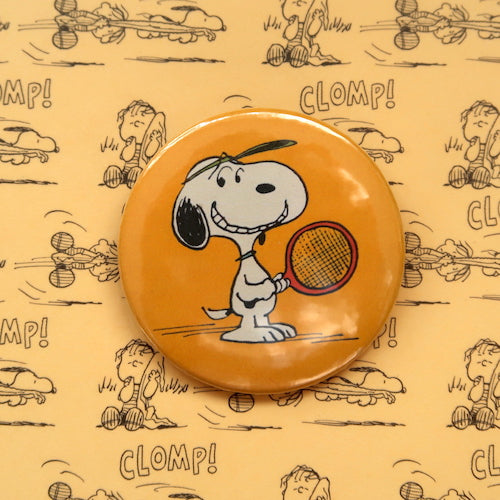 A vintage style pin button badge featuring Snoopy the beagle from the Peanuts comics playing tennis