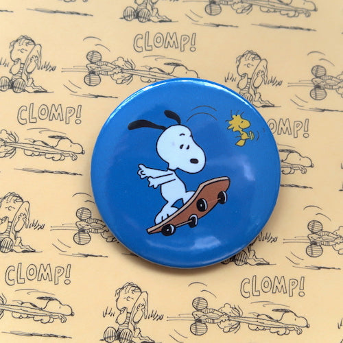 A vintage style pin button badge featuring Snoopy the beagle from the Peanuts comics skateboarding with Woodstock