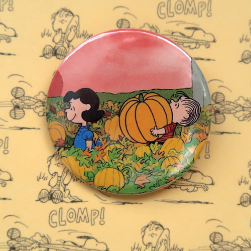 A vintage style pin button badge featuring Linus and Lucy from the Peanuts comics carrying a pumpkin