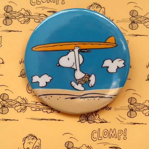 A vintage style pin button badge featuring Snoopy the beagle from the Peanuts comics on the beach with a surfboard