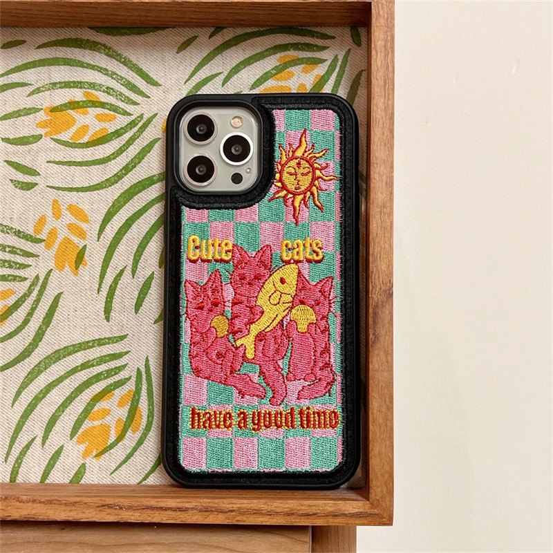 A black embroidered iPone case featuring an embroidered illustration of pink cats with fish and balls and a yellow sun under "cute cats have a good time" lettering slogan
