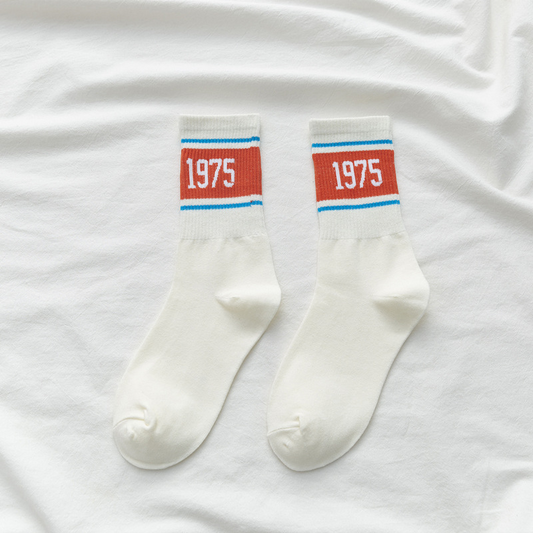 A pair of white crew socks with a red, white, and blue stripe and the number 1975 printed on the ankle area.